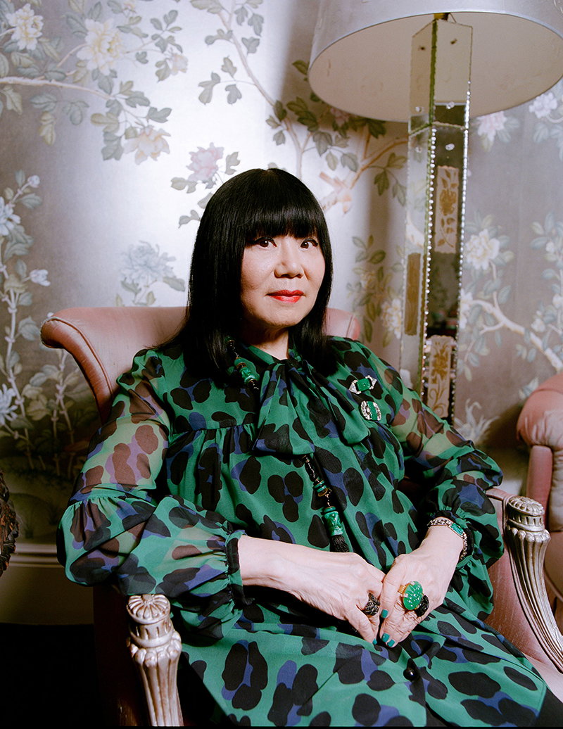 Anna Sui, sitting in a chair and wearing a teal leopard-print blouse.