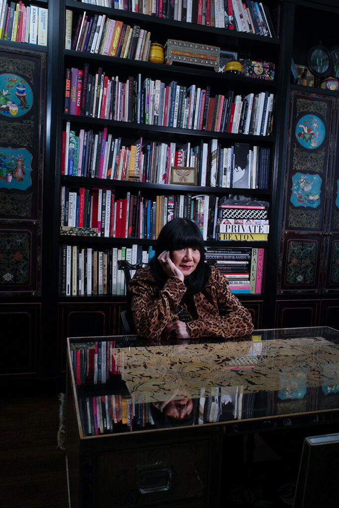 Anna Sui sitting at a desk in front of a large bookshelf.